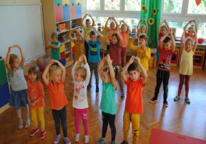 dzieci ubrane na kolorowo ustawione w sali w rzędach śpiewają piosenkę i ilustrują ją ruchem